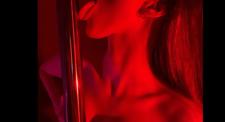 Young VR pole dancer stripper licks the pole with her tongue bathed in red neon lighting.